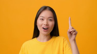 Neşeli bir Asyalı kadın, sanki bir fikri ya da öneriyi paylaşır gibi, canlı sarı bir arkaplanda, gülümsüyor ve parmağını kaldırıyor. Günlük kıyafetleri hayat dolu atmosferi tamamlıyor..