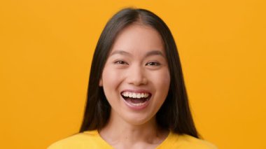 Düz, uzun saçlı genç bir kadın ışıl ışıl gülümsüyor, neşe ve pozitiflik saçıyor. Canlı, sarı bir zemin üzerinde duruyor, neşeli ve canlı bir atmosfer sergiliyor..