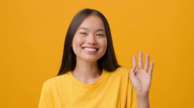 Uzun saçlı genç bir Asyalı kadın ışıl ışıl gülümseyip elini sallıyor. Neşeli sarı bir gömlek giyiyor, neşeli bir atmosfer yaratıyor ve neşe ve pozitifliği yakalıyor..