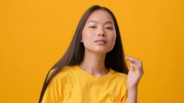 Parlak sarı bir gömlek giymiş Asya kökenli genç bir kadın, çarpıcı sarı bir arkaplana karşı güven içinde poz veriyor. Uzun saçları kamerayla şakalaşırken nazikçe akıyor..