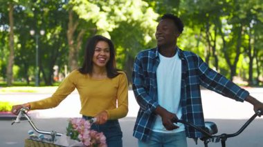 Genç bir Afrikalı Amerikalı çift, yeşil alanlarla dolu canlı bir parkta bisiklet sürüyor, kahkaha ve neşe paylaşıyorlar. Kadın elinde bir buket çiçek tutuyor, neşeli atmosferi yükseltiyor..