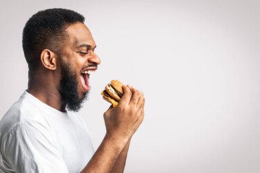Beyaz arka planda duran, stüdyoda çekim yapan, hamburger yiyen aç siyah adamın yan görüntüsü. Metnin için boş yer. Abur cubur, sağlıksız erkek beslenme ve aşırı yeme alışkanlığı..