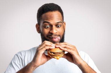 Beyaz Stüdyo Arkaplanında Lezzetli Burger Duran ve Dudaklarını Yalayan Aç Siyah Adam. Sağlıksız beslenme ve abur cubur yeme alışkanlığı, aldatma yemeği konsepti