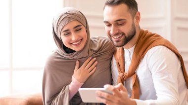 Neşeli Müslüman çift akıllı telefon kullanıyor cep telefonlarında komik videolar izliyorlar evde koltukta birlikte otururken eğleniyorlar. İnsanlar ve Modern Gadget 'lar. Cep Telefonu için Yeni Uygulama