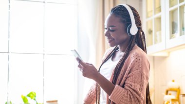 Akıcı Uygulama. Kablosuz kulaklıkla neşeli siyah kadın portresi rahat mutfağın içinde akıllı telefonlardan müzik dinliyor ve evde dinlenirken sosyal ağları tarıyor.