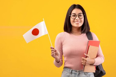 Dil eğitimi ve değişim öğrencisi. Sırt çantası ve kitaplarıyla, elinde Japon bayrağı, turuncu arka planda izole edilmiş, fotokopi alanı, stüdyo çekimi...
