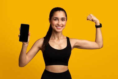 Egzersiz Başvurusu. Spor kıyafetleri içinde gülümseyen genç bir kadının portresi. Elinde cep telefonu olan, şablon için siyah, boş bir ekranla turuncu stüdyo arka planında izole edilmiş pazıları gösteren bir resim.