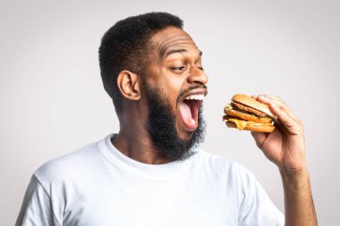 Beyaz arka planda stüdyoda durup hamburger yiyen Afro-Amerikalı adamın yan görüntüsü. Abur cubur ve aşırı yeme alışkanlığı. Zenci Isıran Çizburger Sağlıksız Beslenme Konsepti