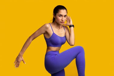 Yüksek Yoğunluklu Ara Eğitim. Kendine güvenen genç bir kadının portresi bacağını kaldırıyor, kolunu kaldırıyor, diz kapağı egzersizi yapıyor turuncu stüdyoda tek başına ayakta duruyor.