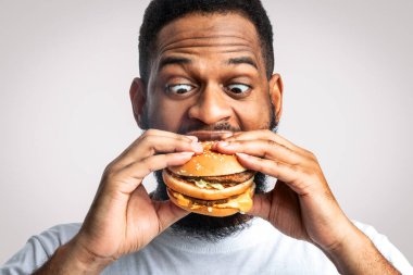 Beyaz Stüdyo Arkaplanında Burger Yiyen Komik Afrikalı Adam. Siyah adam hamburgerin tadını çıkarıyor, abur cubur yiyor. Beslenme ve Sağlıksız Fast food, Aşırı yeme alışkanlığı sorunu