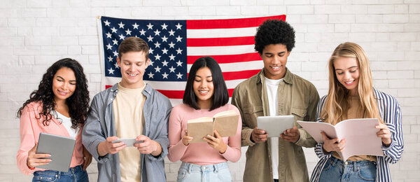 Modern study in USA, language learning and student exchange program. Cheerful busy young international people reading books and gadgets on white wall background with large flag of America, studio shot