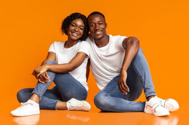 Kaygısız Afrikalı Amerikalı aşıklar yerde oturup sarılıyorlar, turuncu arka plandaki kameraya gülümsüyorlar, stüdyo görüntüleri, fotokopi çekimleri. Neşeli bin yıllık siyah erkek ve kadın birlikte poz veriyor.