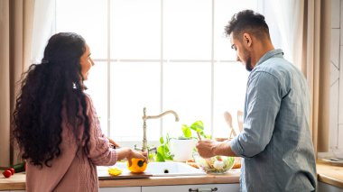 Mutfak penceresinin yanında birlikte yemek hazırlayan mutlu Orta Doğulu çift, Gülümseyen Arap adam ve kadın öğle yemeği için sağlıklı sebze yemeği pişiriyor, vejetaryen mutfağının tadını çıkarıyor,