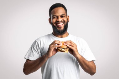 Abur cubur. Stüdyoda beyaz arka planda durup kameraya gülümseyen mutlu siyah adam hamburgeri tutuyor. Beslenme, aşırı yeme alışkanlığı ve sağlıksız yeme anlayışı