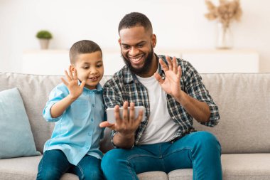 Afro-Amerikan Baba ve Oğlu, telefonlarıyla evdeki koltukta oturan akıllı telefon ekranına el sallayarak selam veriyor. Baba ve Çocuk Çevrimiçi İletişim Kuruyor. Uzaktan iletişim. Seçici Odaklanma