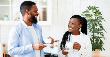 Çiftler Günlük Hayatı. Mutfakta birlikte sabah kahvesi içen mutlu siyah adam ve kadın, genç Afrikalı Amerikalı aşıklar sıcak içeceklerle bardaklarını tutuyorlar ve birbirlerine gülümsüyorlar, hafta sonunun tadını çıkarıyorlar.