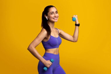 Güç Antrenmanı Konsepti. Gülümseyen Fit Bayan 'ın portresi, iki dambılla egzersiz yapmak, kablosuz kulaklık takmak, portakal rengi stüdyo arka planında izole edilmiş pozlar vermek, egzersiz yapmak, kameraya bakmak.