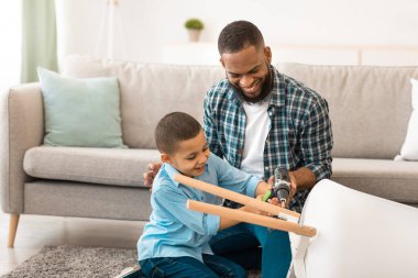Afrika kökenli Amerikalı baba ve oğul evde ev işleri yaparken elektrikli matkabı kullanarak masayı tamir ediyorlar. Mutlu Çocuk, Ev İşi Konseptinde Babaya Yardım Ediyor. Seçici Odaklanma