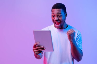 Çevrimiçi Kazanan. Afro-Amerikalı neşeli adam dijital tabletle başarıyı kutluyor, duygusal mutlu siyah adam yumruklarını kaldırıyor ve heyecanla haykırıyor, mor arka planda neon ışığı altında duruyor.