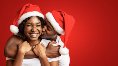 Noel Baba şapkalı romantik siyah adam arkadan sarılıyor ve gülüşen kız arkadaşını öpüyor, turuncu stüdyo arka planı, yakın plan. Afro-Amerikan çiftleri birbirlerini seviyorlar, yeni yılı kutluyorlar