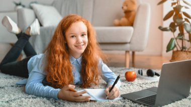 Çevrimiçi Eğitim. Kırmızı saçlı genç kız dizüstü bilgisayarı ve not defteriyle yerde yatıyor. Öğrenci ders veya web sitesi izliyor, evde çalışıyor, interneti kullanıyor, kameraya bakıyor.