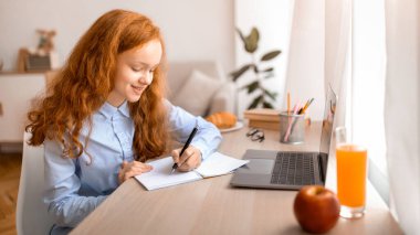 Uzaktan eğitim konsepti. Gülümseyen genç kız masada oturuyor, dizüstü bilgisayar kullanıyor ve defterine ya da günlüğüne bir şeyler yazıyor. Öğrenci kız çevrimiçi kurs izliyor, not alıyor, ödev yapıyor.