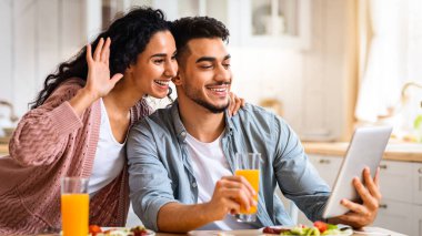 Neşeli Arap eşler, mutfakta kahvaltı yaparken dijital tablete video çağrısı yapıyor, gülümseyen Orta Doğu çifti, web kamerasına el sallıyor, birini selamlıyor, çevrimiçi iletişimin tadını çıkarıyor.