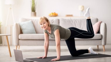 Çevrimiçi ev sporları konsepti. Oturma odasında dizüstü bilgisayarın önünde spor yapan son sınıf atletik bir kadın. Yetişkin bir kadın ev içi spor yapıyor, yoga minderi üzerinde egzersiz yapıyor, kaslarını güçlendiriyor.