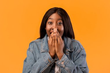 Kot ceketli, ağzı turuncu stüdyo arka planı, yakın plan, fotokopi alanı ile kaplanmış siyahi bir kadın. Afrikalı Amerikalı genç bir kadın şok edici ya da heyecan verici bir şey görüyor.