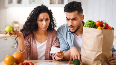 Şok edici Fiyatlar. Market alışverişinden sonra faturaları kontrol eden kafası karışmış Orta Doğulu eşler, mutfakta oturan genç Arap çift, süpermarket faturalarını okuyan üzgün adam ve kadın,