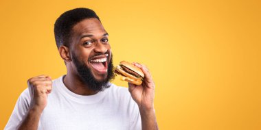 Heyecanlı Afrikalı Adam Burger Yiyor Evet, Sağlıksız Fast Food 'un tadını çıkarıyor Sarı Stüdyo Arkaplanının Üzerinde Duruyor. Son olarak, Hileci Yemek. Lezzetli Atıştırmalık Yiyecek Konsepti
