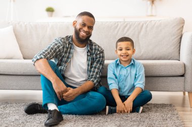 Siyah Baba ve Oğlu, evdeki oturma odasında yerde oturmuş kameraya gülümsüyor. Neşeli bir baba ve çocuğunun portresi. Aile bağı kavramı
