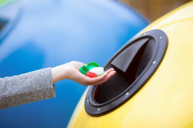 Tanınmayan bir kadın, bir avuç dolusu renkli plastik kapağı sarı geri dönüşüm kutusuna atıyor, çöp kutusunun keyfini çıkarıyor ve hiç atık ayıklamıyor, seçici odaklı kesilmiş resim, boş alan...