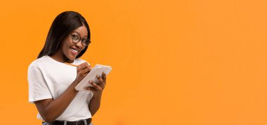 Gözlüklü, gülümseyen ve not alan, elinde not defteri ve kalem tutan, turuncu arka plan, kopyalama alanı olan zeki siyahi bir kadın. Neşeli zeki Afro-Amerikan Kadın Planlama Günü