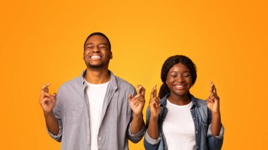 Bir dilek tut. Batıl inançlı siyah çift parmakları çapraz ve gözleri kapalı Afrika kökenli Amerikalı erkek ve kadın, sarı arka planda yan yana dururken şans dileniyor.