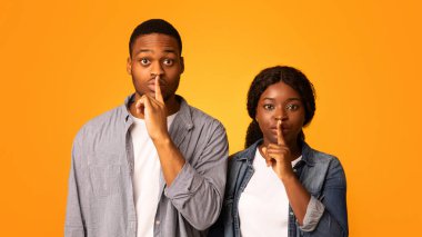 Tek kelime etme. Siyahi adam ve kadın parmaklarını dudaklarında tutuyor, sessiz işareti yapıyor ve kameraya bakarken sessiz bir jest yapıyor, genç Afrikalı çift sarı arka planda yalnız duruyor.