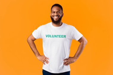 Neşeli Afrikalı gönüllü adam, elleri kalçasında, gönüllü olarak çalışmaya hazır, sarı stüdyoda poz veriyor, kameraya gülümsüyor. Gönüllü İş, Eylemciler Portresi