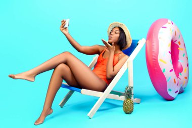 Flört eden siyahi kadın, erkek arkadaşıyla akıllı telefondan sohbet ediyor, webcam 'de canlı yayında öpüşüyor, plajda şezlongda otururken selfie çekiyor, mavi stüdyo arka planında. Tam boy portre