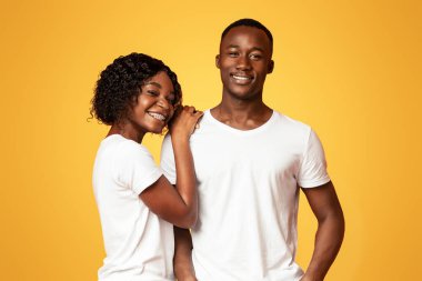 Turuncu stüdyo arka planında birlikte poz veren, kucaklaşan ve gülümseyen kot pantolonlu ve beyaz tişörtlü çekici erkek ve kadın. Aşk, aile, ilişkiler