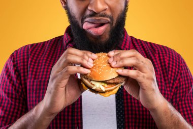 Aç Afro-Amerikalı Adam Burger yiyor ve dudaklarını yalıyor. Sarı Stüdyo Arkaplanı Üzerinde Lezzetli Çizburger Pozu Veriyor. Abur cubur yeme alışkanlığı, sağlıksız beslenme ve aldatma yemeği.