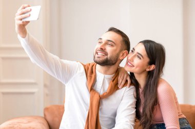 Neşeli bir Arap çift cep telefonuyla selfie çekiyor evdeki koltukta oturuyor. Modern Orta Doğu Milenyumu 'nun eğlenceli pozları ve kapalı mekanda cep telefonuyla fotoğraf çekimleri. İnsanlar ve Gadget