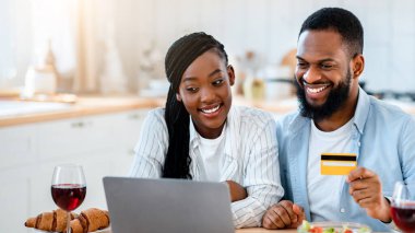 Yemek siparişi. Mutlu siyah eşler mutfakta dizüstü bilgisayar ve kart kullanıyor, bilgisayarla online teslimat siparişi veriyor, masada oturuyor ve internet alışverişlerinin tadını çıkarıyor,