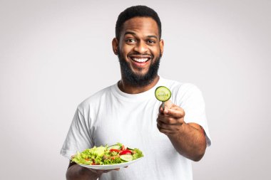 Sağlıklı beslenme. Sebze salatasıyla poz veren neşeli Afrikalı Adam Beyaz Stüdyo Arkaplanının başında duran kameraya salatalık dilimi sunuyor. Vegan Diyet, Erkek Beslenme ve Kilo Verme Konsepti 'ni deneyin.