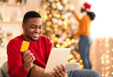Modern teknoloji ve hızlı hediye alışverişi. Şaşırmış bin yıllık Afro-Amerikan erkeği, tablete bakıyor ve kredi kartı tutuyor, kadın Noel ağacını süslüyor parlayan çelenkler, kopyalama alanı