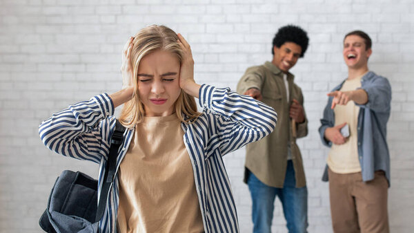 Bullying and peer relationships. Focus on blonde teen lady with backpack covering her ears, not to hear taunts from multiracial male students in school or college, on white brick wall background
