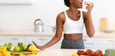 Siyah Fitness Kadınları Evdeki Mutfakta Aldatma Yemekleri Yiyen Donut Poz Veriyor. Şekerli yiyecekler ve tatlılar, kötü beslenme alışkanlığı, abur cubur arzusu ve aşırı yeme kavramı.