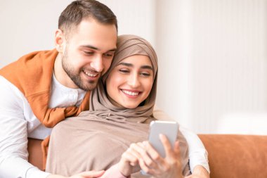 Mutlu Müslüman çift akıllı telefon mesajları kullanıyor ve evdeki koltukta oturup interneti inceliyor. Aile eğlencesi. Neşeli Arap Eşler İnternette Video İzliyor. Büyük Mobil Uygulama Konsepti