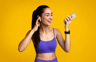Spor için müzik. Kablosuz kulaklık takan gülümseyen genç bayanın portresi, en sevdiği şarkı ve müzik listesinin keyfini çıkartıyor, akıllı telefonu portakal rengi stüdyonun arka planında izole halde tutuyor.