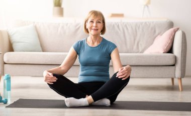 Lotus 'ta oturmuş spor minderinde poz veren, evde meditasyon yapan, fotokopi çeken yaşlı bir kadın. Yoga yapan dengeli olgun bir bayan, iç huzurunu ve farkındalığını buluyor.