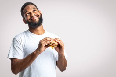 Hile Yemeği. Mutlu Siyahi Milenyum Adamı Lezzetli Burger Beyaz Stüdyo Arkaplanının Üzerinde Rüya Gibi Duruyor. Abur cubur yiyici konsepti. Beslenme ve aşırı yeme alışkanlığı